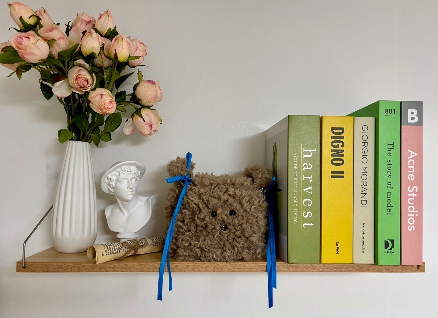 A shelf with a vase of pink roses, a white bust, a teddy bear, and a stack of books including titles like "DIGNO II" and "harvest".