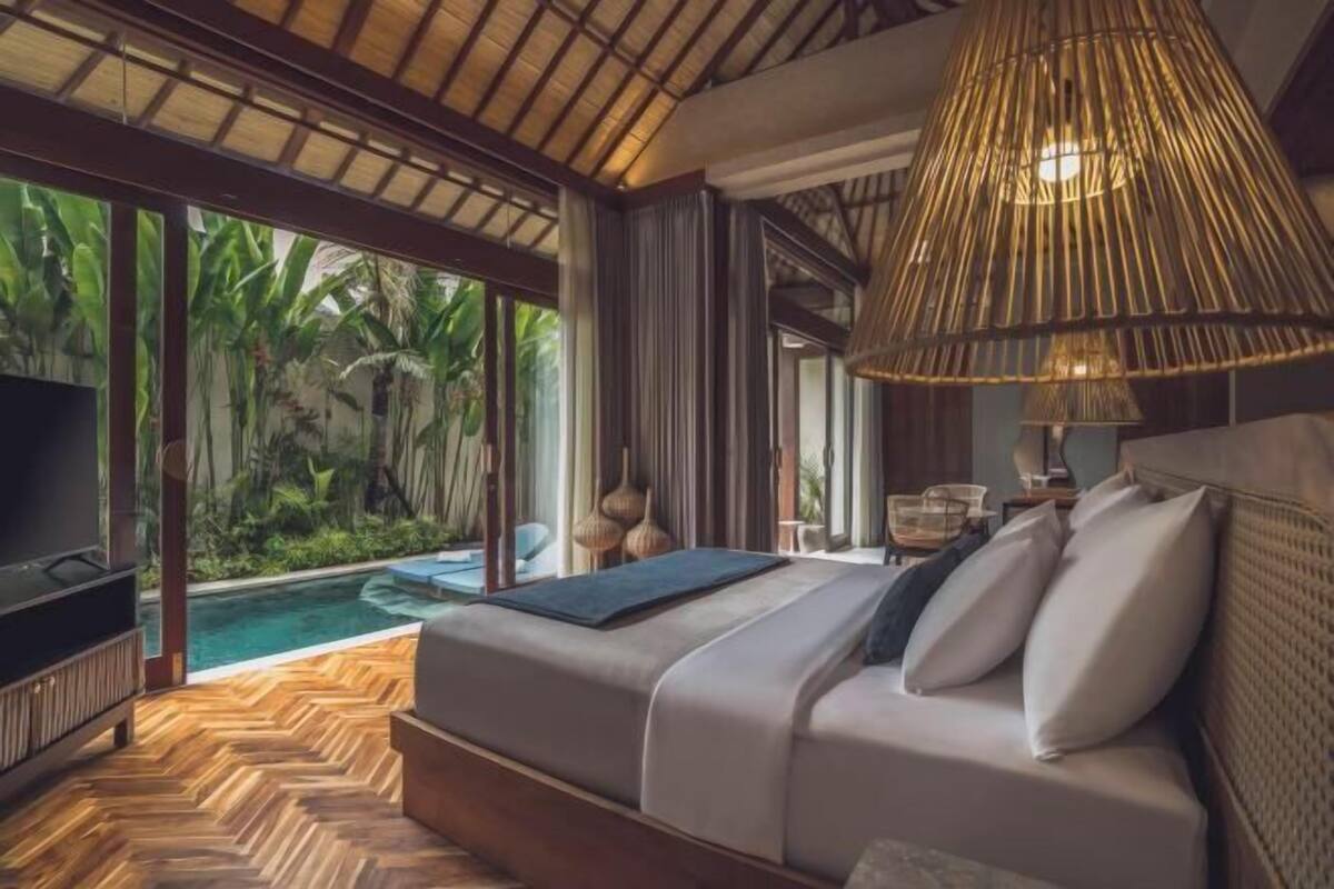 A bedroom with a bed, a TV, a large window, and a hanging light fixture.