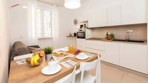 A modern kitchen with white cabinets, a wooden dining table, and a breakfast setting with fruits and pastries.