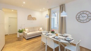 Una sala de estar moderna con sofá, mesa de comedor y sillas. Se observa un reloj de pared grande y un adorno decorativo.