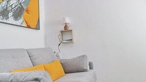 Un sofá gris con un cojín amarillo, un cuadro en la pared y una mesita con una lámpara.