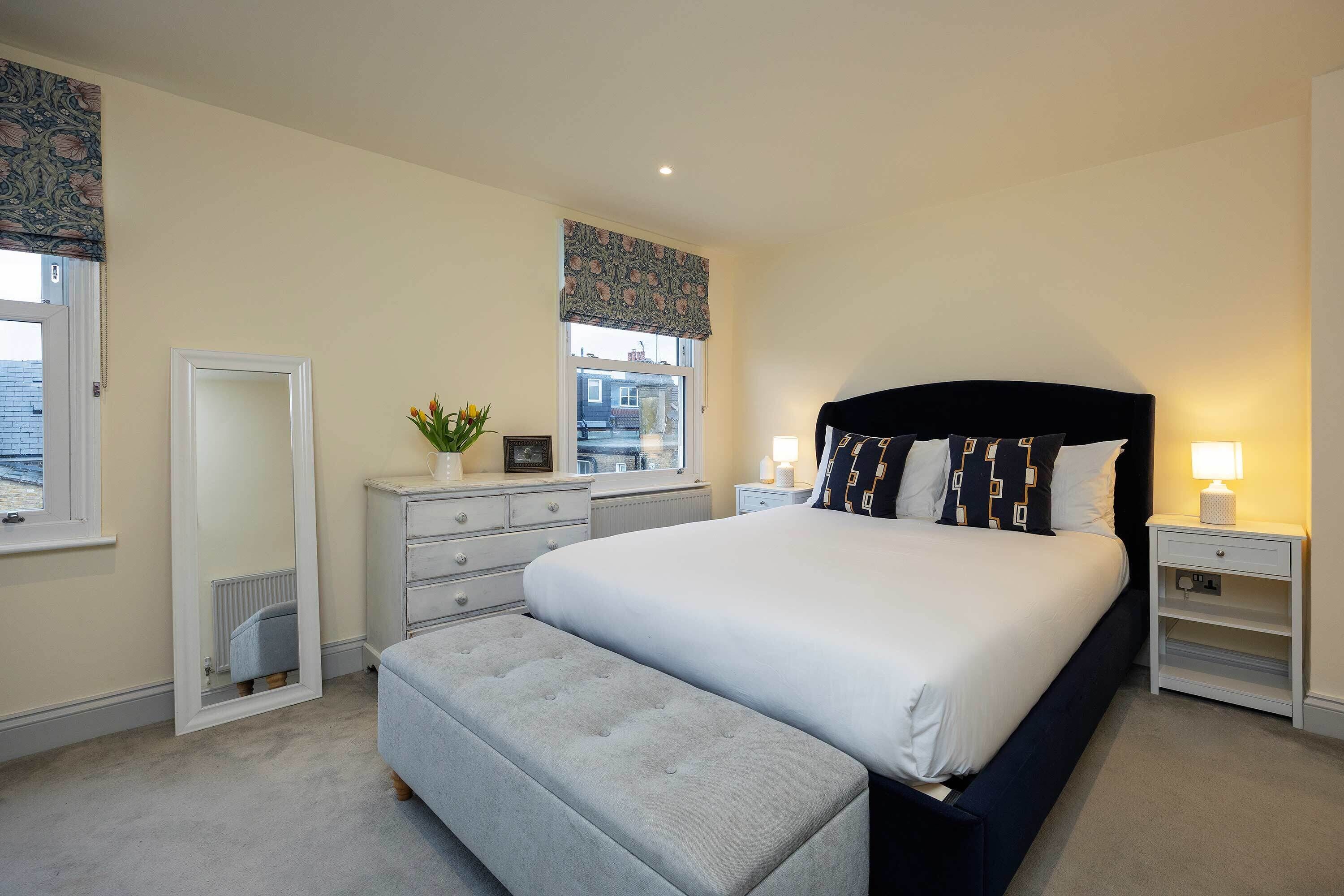 A bedroom with a large bed, a bedside table with a lamp, a dresser, and a window with curtains.