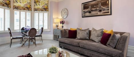A living room with a brown sofa, a dining table with chairs, a coffee table with a tray, and a clock on the wall.