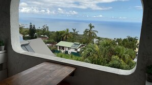 A room with a view of the ocean, a wooden table, and a window with a concrete frame. - 15 minutes from the beaches of St Leu or Étang Salé. Sea view apartment with swimming pool. (Saint-Leu)