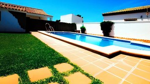 Piscina com um deck ladrilhado, rodeada por um espaço gramado e uma parede branca.