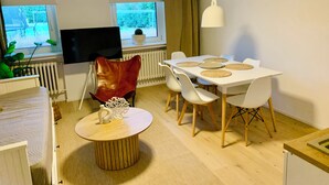 A modern living room with a dining area, a flat-screen TV, and a view of greenery outside.