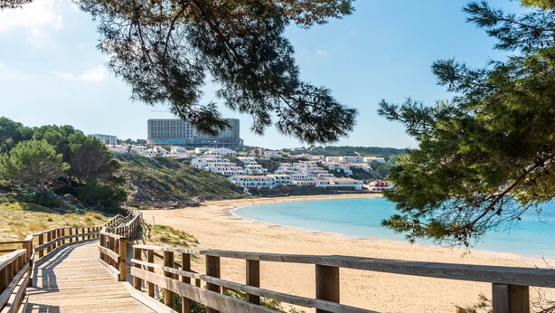 A wooden boardwalk leading to a beach with a view of a coastal town and a large building in the background. - CASTELLSOL 201 (Arenal d´en Castell)