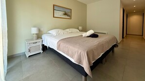 A hotel room with a large bed, a nightstand, a lamp, a painting on the wall, and a hallway leading to another room.
