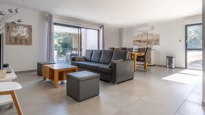 A modern living room with a grey sofa, wooden coffee table, and ottoman. There is a dining area with a table and chairs, and a large window providing natural light.