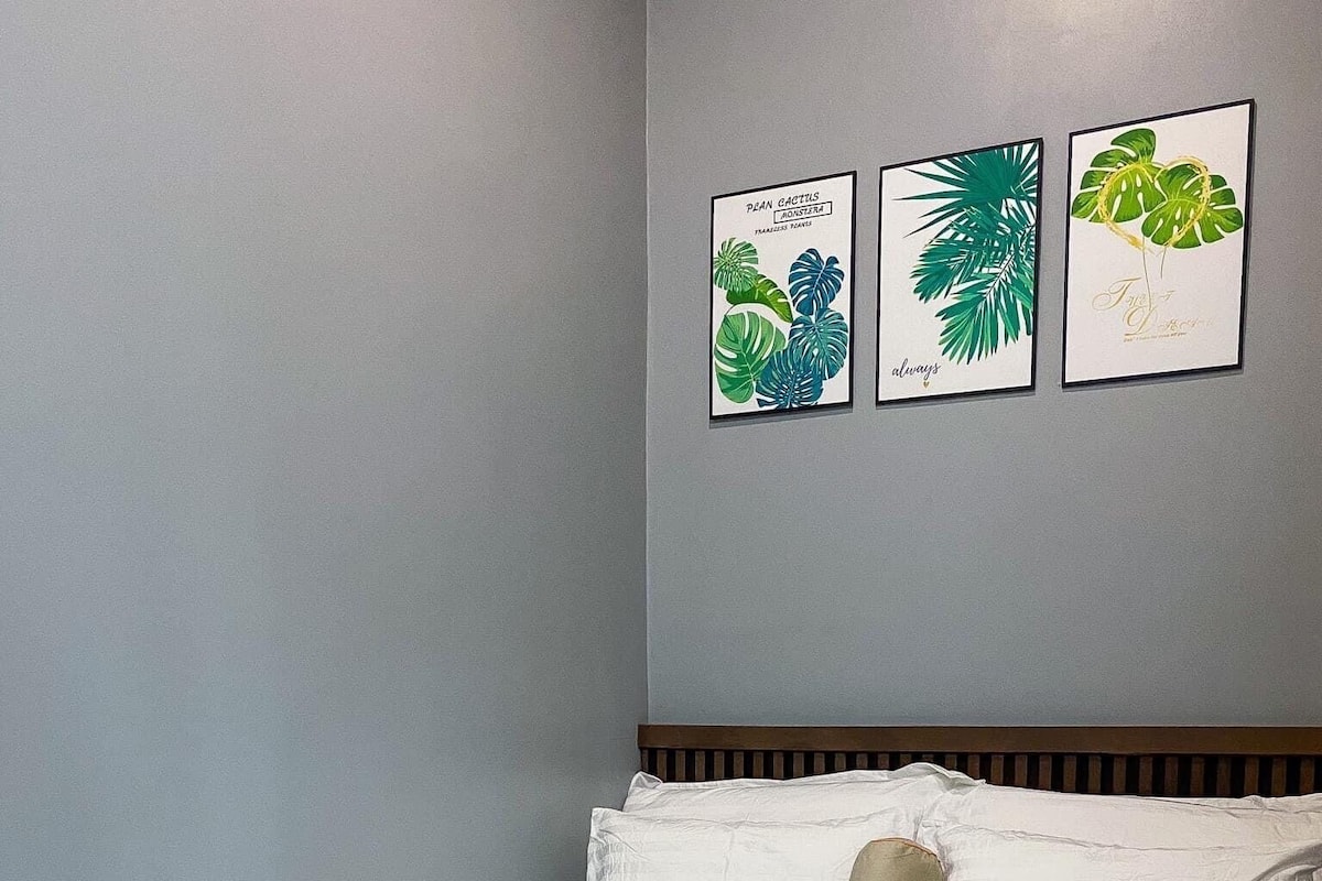A neatly made bed with white bedding in a room with light gray walls. There are three framed botanical prints on the wall above the bed.