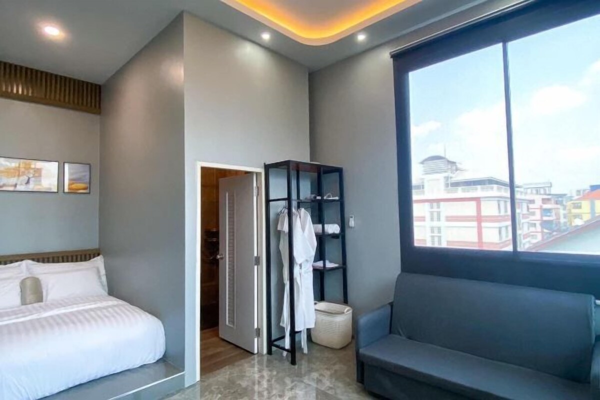 A modern hotel room with a bed, a seating area, a wardrobe, and a view of buildings.