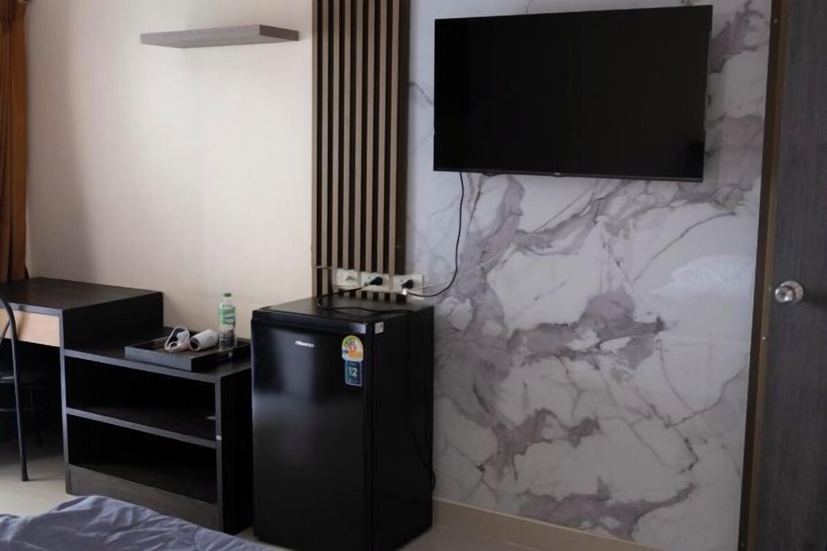A hotel room with a bed, a desk with a chair, a black minibar, a TV mounted on the wall, and a marble-patterned wall.