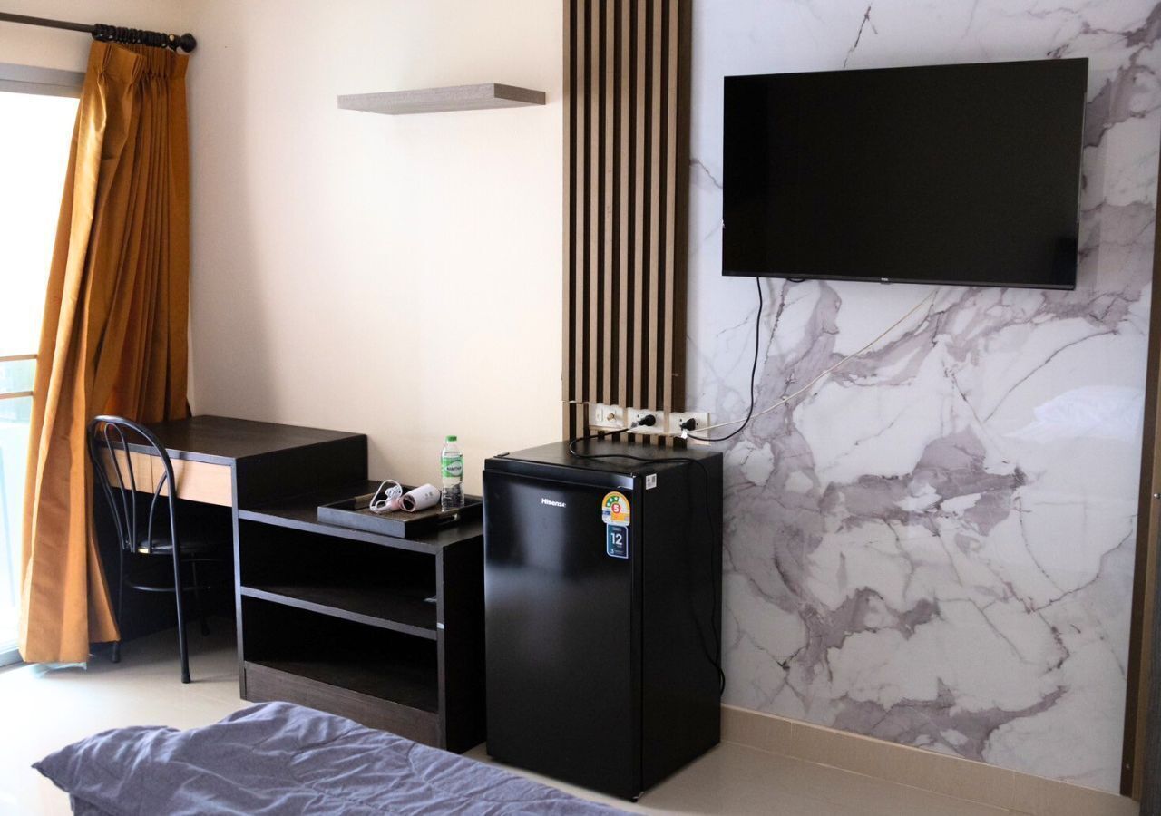 Una camera compatta con un letto, una scrivania con una sedia, un frigorifero nero, una TV a parete e una finestra con tende.