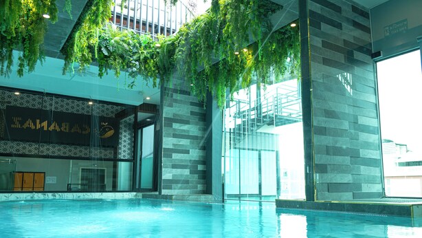 An indoor swimming pool with hanging plants, a patterned wall, and a glass door leading to a staircase. - Cabanaz Pool Villa (Bangkok)