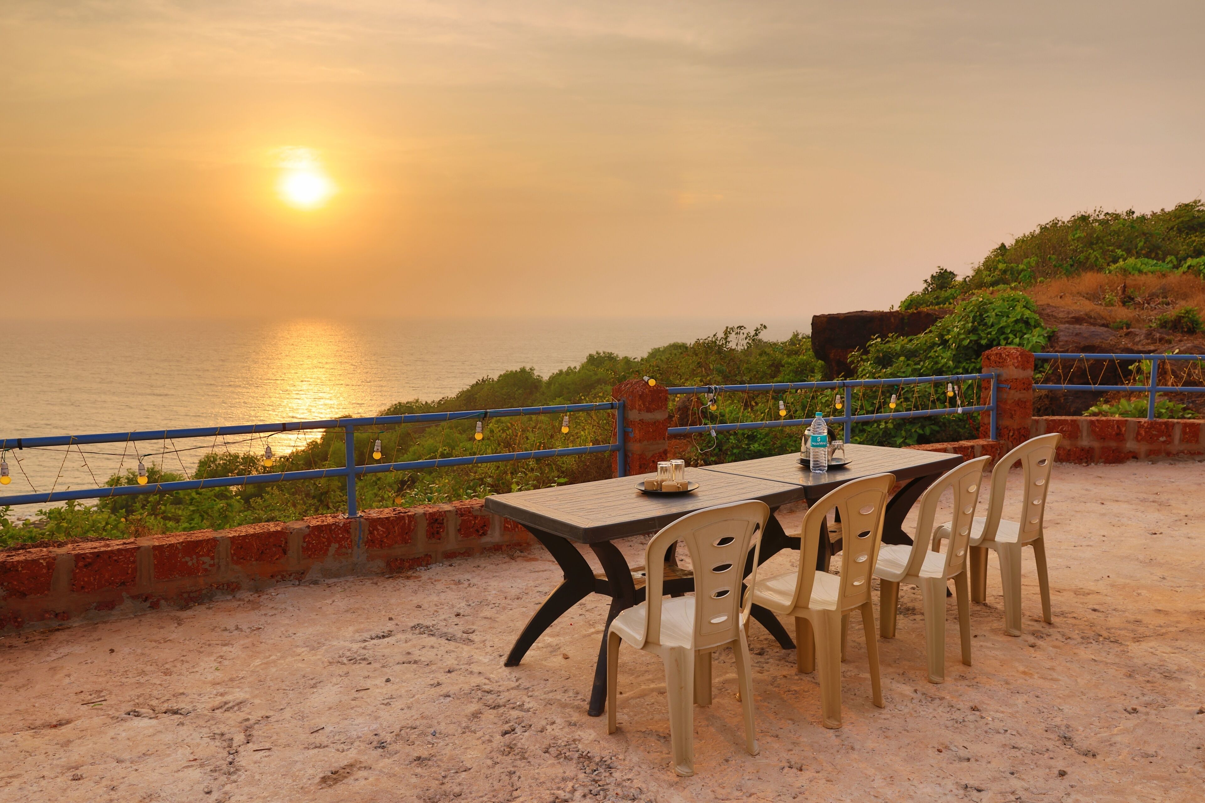 A table with chairs set up on a sandy surface, overlooking a body of water during sunset.