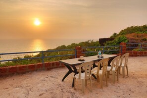 Une table entourée de chaises, installée sur une surface sablonneuse, d’où l’on peut admirer un plan d’eau au coucher du soleil.