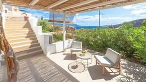 A covered outdoor seating area with a staircase leading to a terrace, surrounded by greenery and overlooking a scenic landscape. - Psathaki  (Kimolos)