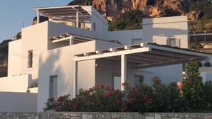 A modern white building with a stone wall in front, balconies, and red flowers. - Psathaki  (Kimolos)