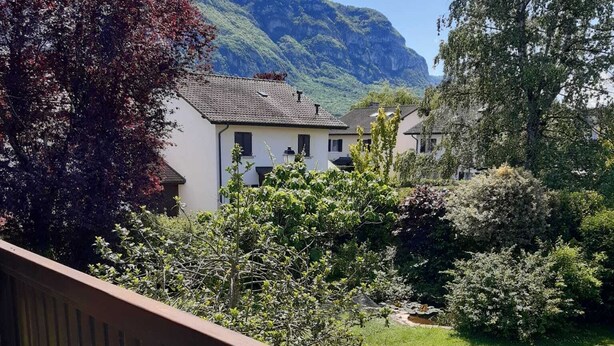 Un balcon avec une table, une bouteille de vin et un verre, donnant sur un jardin luxuriant et des maisons, avec une chaîne de montagnes en toile de fond.