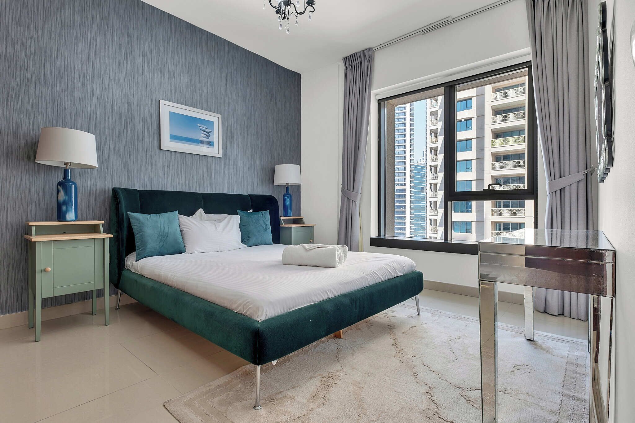 A modern bedroom with a large bed, two bedside tables, a nightstand, a glass coffee table, and a view of high-rise buildings.