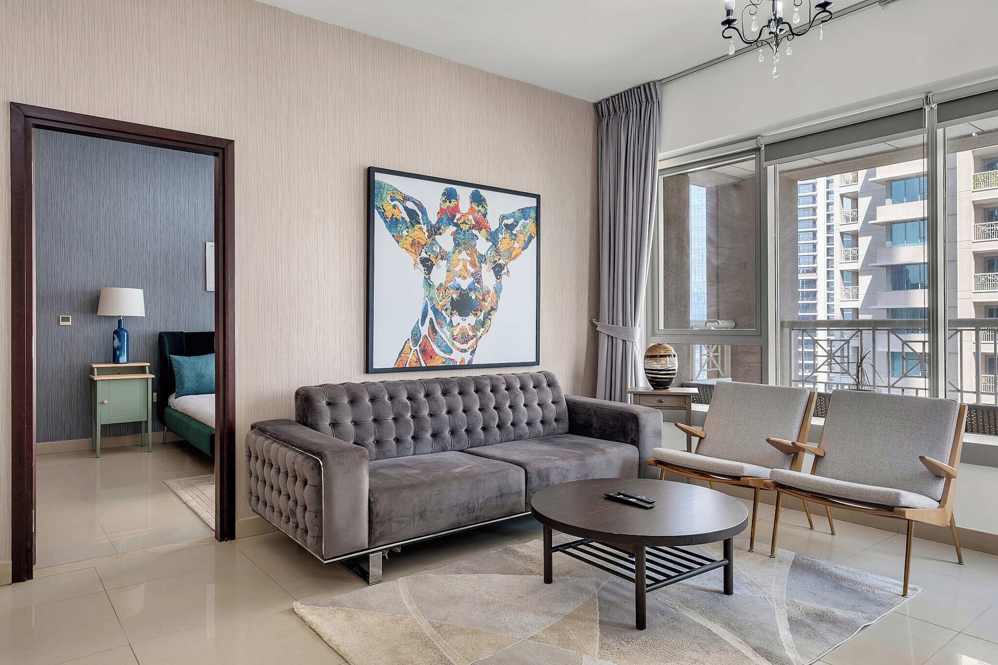 Un salon moderne avec un canapé gris, deux fauteuils, une table basse et une grande fenêtre donnant sur des immeubles.