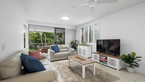 A modern living room with a sofa, coffee table, TV, and a balcony with a view of greenery. - Drift at Casuarina, Pool View - 14 (Tweed)