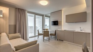 Una habitación de hotel moderna con sofá, mesa de comedor, sillas, televisión y una zona de cocina con armarios y lavamanos.