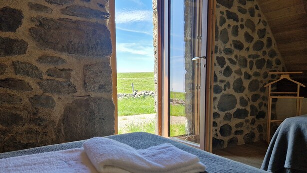 A room with stone walls, a bed with white towels, and a view of a grassy field through a glass door.