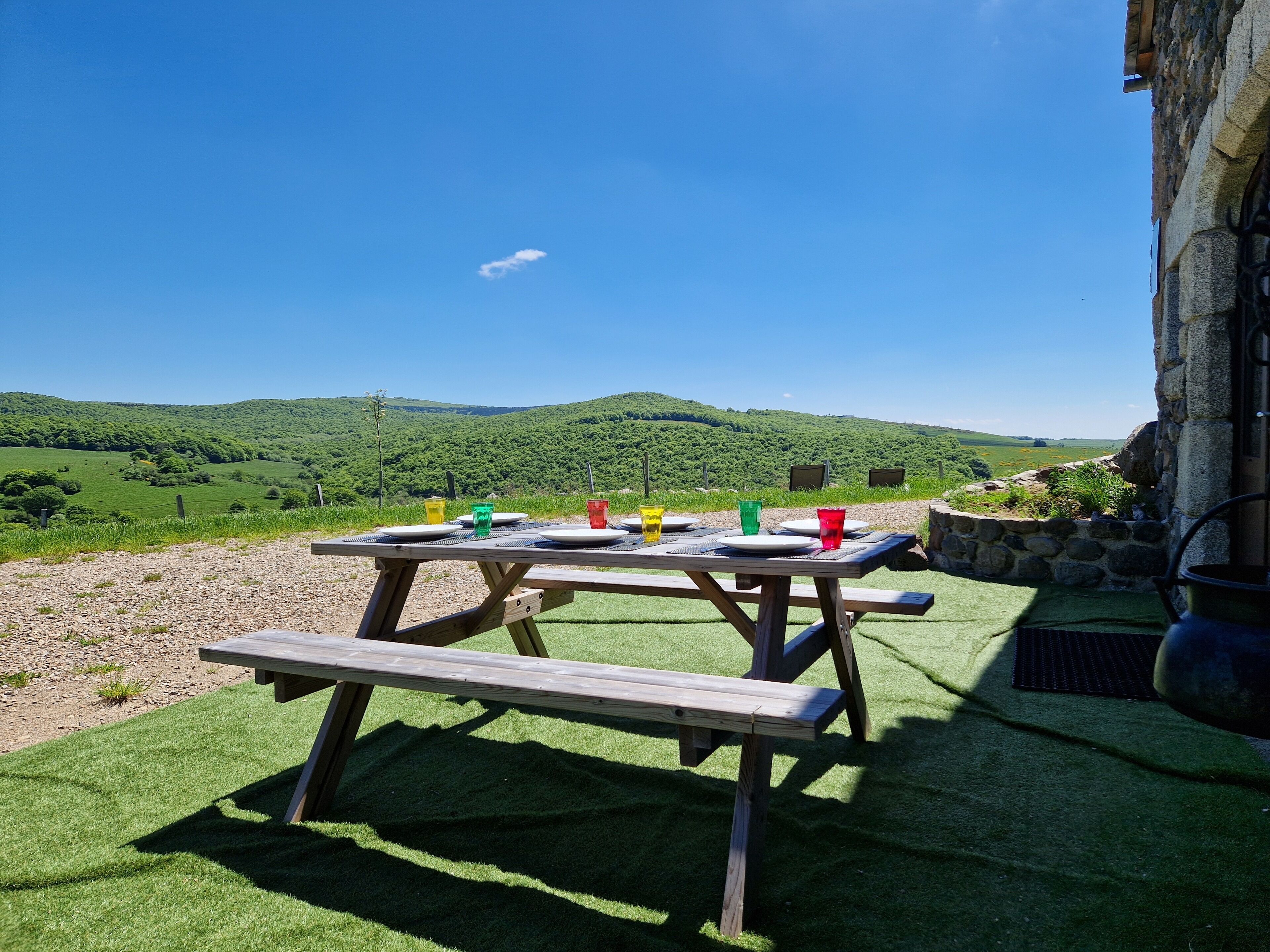 Un tavolo da picnic in legno allestito su un'area erbosa con vista panoramica su dolci colline verdi e un cielo azzurro terso.