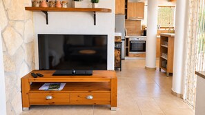 A living room with a flat-screen TV, wooden entertainment unit, stone wall, and a kitchen in the background. - Family Home Iskra (Bulic)