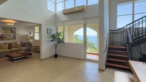 Amplia sala de estar con techo alto, ventanas grandes, una escalera y vista al mar.