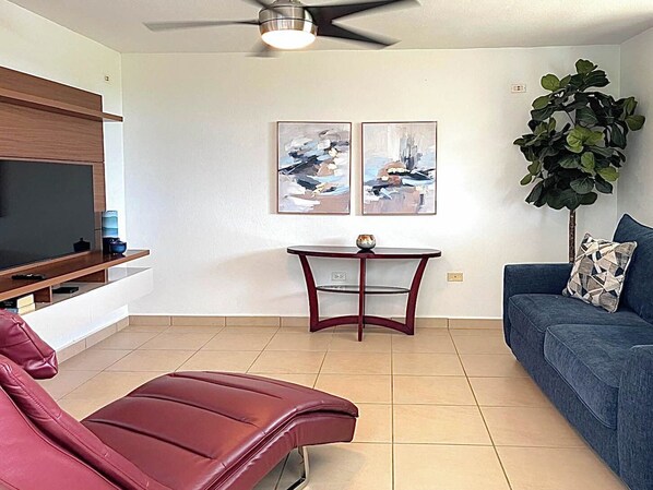 A living room with a TV, a red leather chair, a blue sofa, a small table, and two abstract paintings.