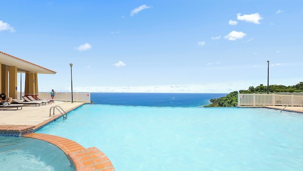 A swimming pool with clear blue water, a tiled deck, and a view of the ocean.