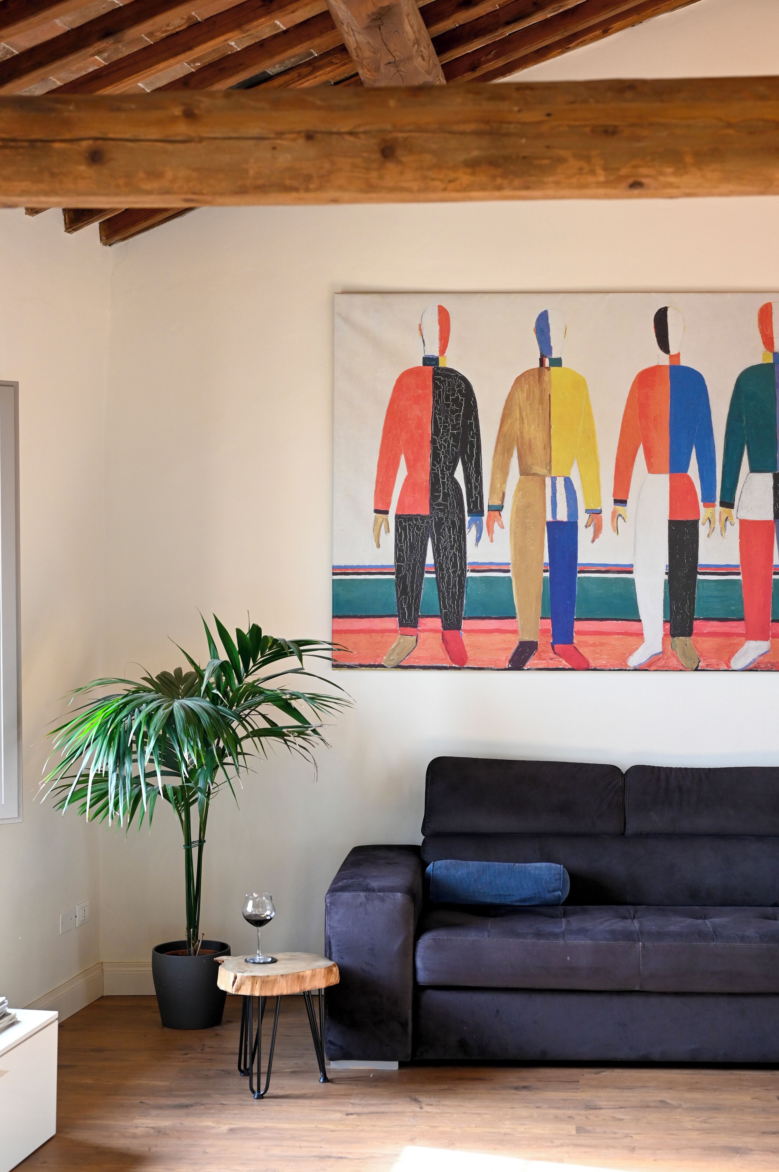 A living room with a dark sofa, a small wooden table with a glass of wine, a potted plant, and a colorful abstract painting on the wall.