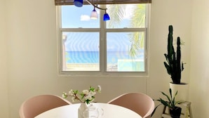Zona pranzo con tavolo rotondo, due sedie, un vaso con fiori e una caraffa. Si vede una finestra con vista su palme e un edificio.
