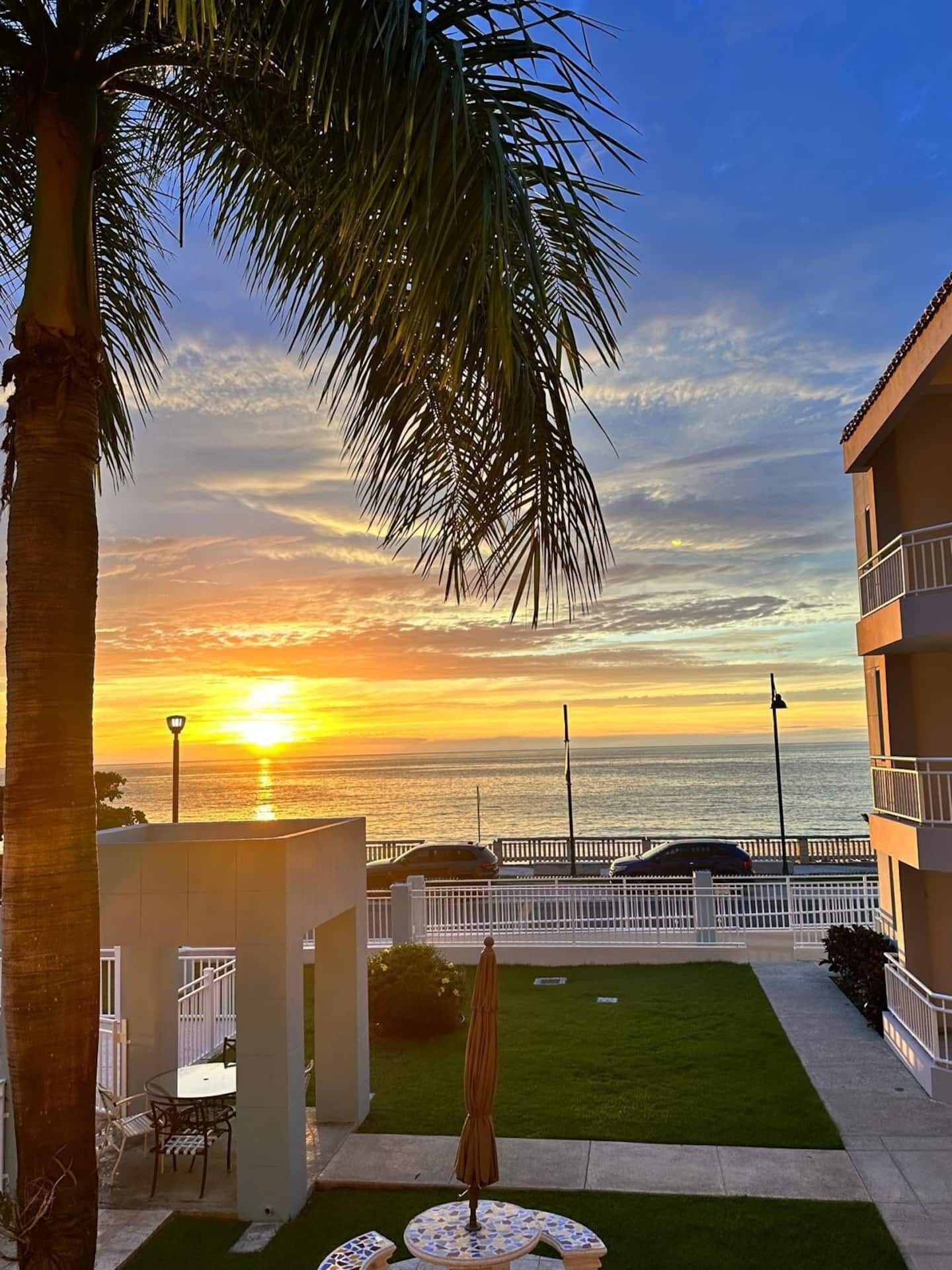 Une terrasse avec une table et des chaises, un petit jardin et une vue sur le coucher de soleil sur l’océan.