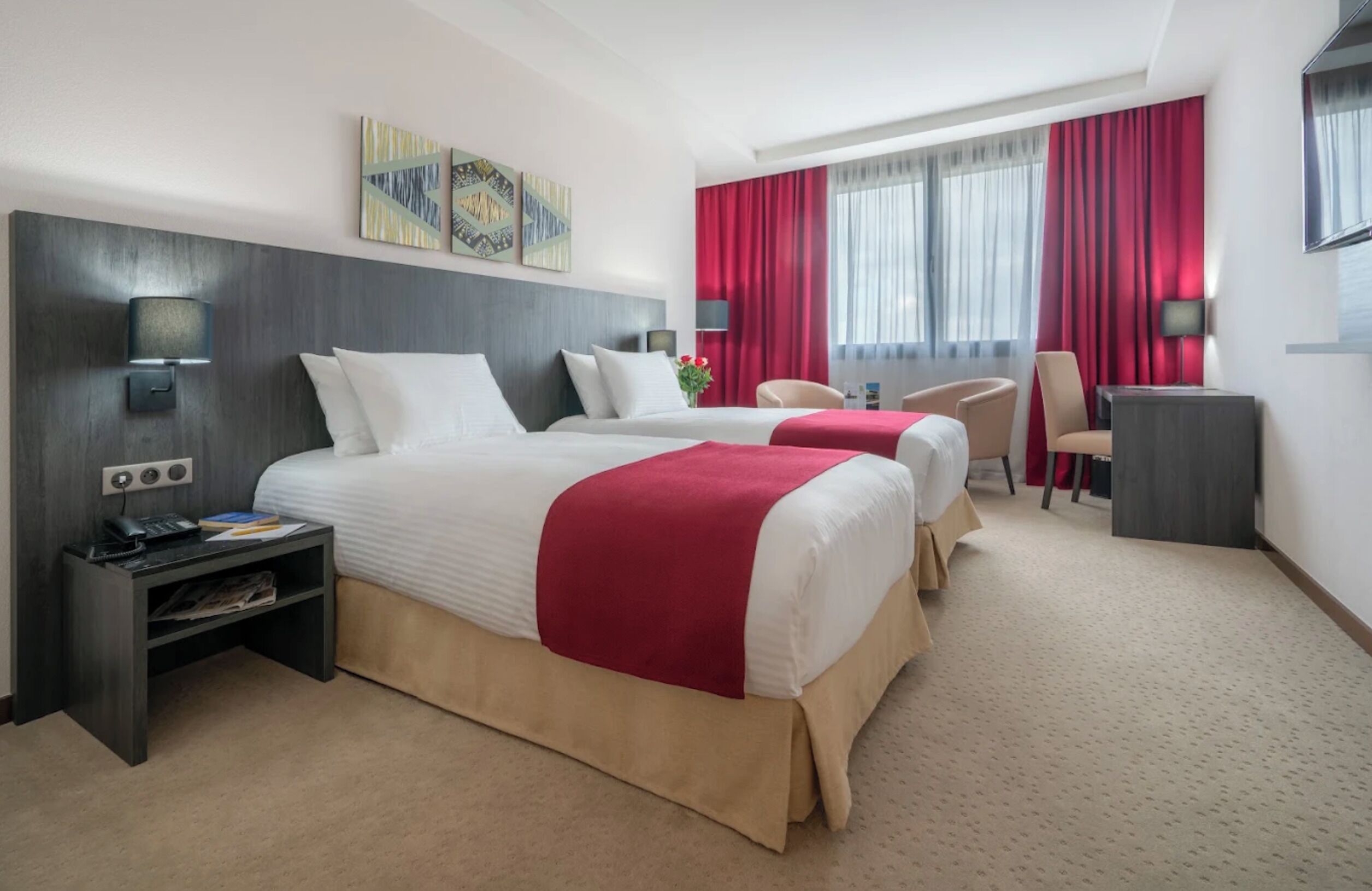 Een hotelkamer met twee bedden, een bureau, een stoel en een raam met rode gordijnen.