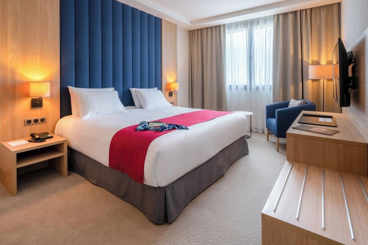 A hotel room with a large bed, two bedside tables, a desk with a chair, a TV, and a window with curtains.