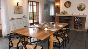 Un coin repas avec une table en bois dressée pour six personnes, entourée de chaises noires à assise tissée. On aperçoit au fond une cheminée agrémentée d’un paravent décoratif.