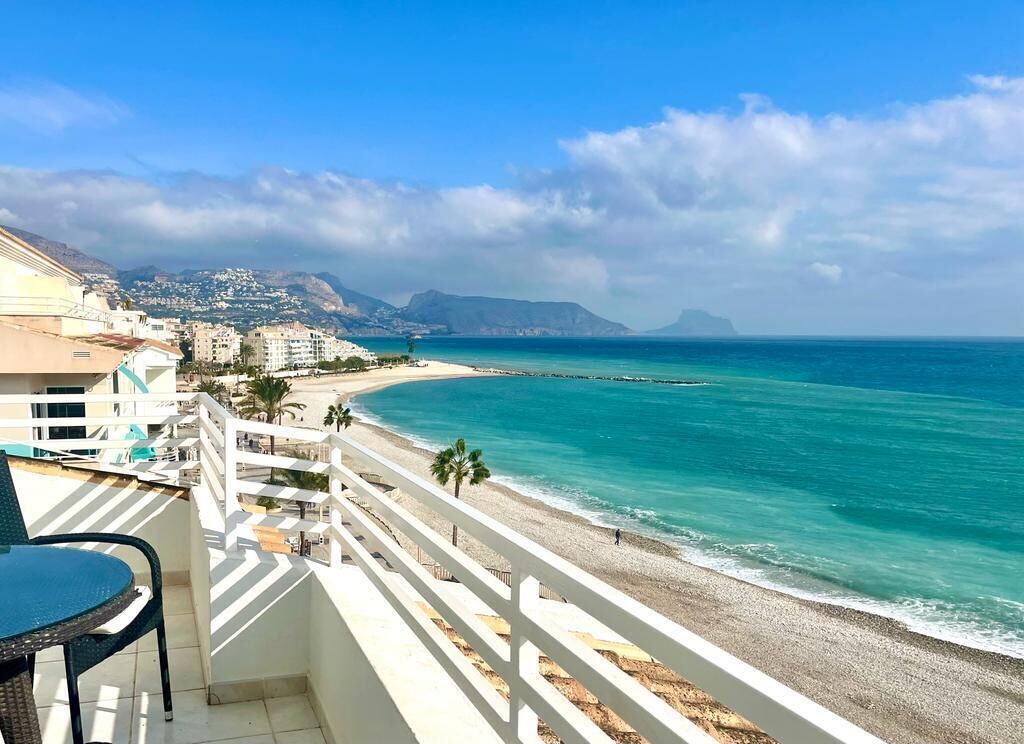A balcony with a view of a beach and ocean, white railings, and a chair.