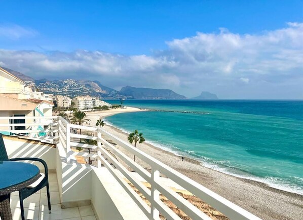 A balcony with a view of a beach and ocean, white railings, and a chair.
