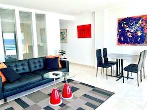 A modern living room with a black sofa, glass coffee table, and red floor lamps. There is a dining area with a white table and black chairs.