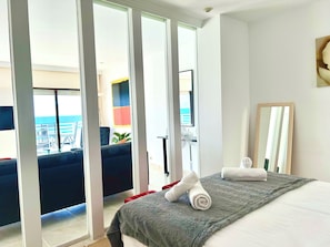 A hotel room with a bed, a large window, a balcony with a view of the sea, and a fan.