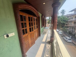 Un balcón con puerta y ventanas de madera, un enchufe, y vista a una calle con coches estacionados y edificios.