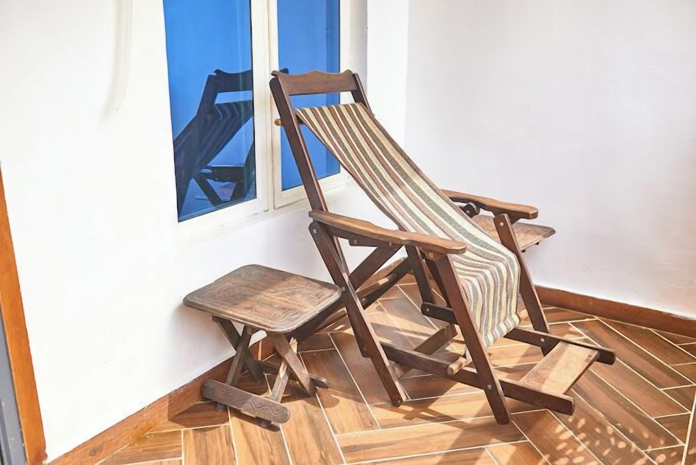 A wooden lounge chair with a striped cushion, positioned on a tiled floor near a window.