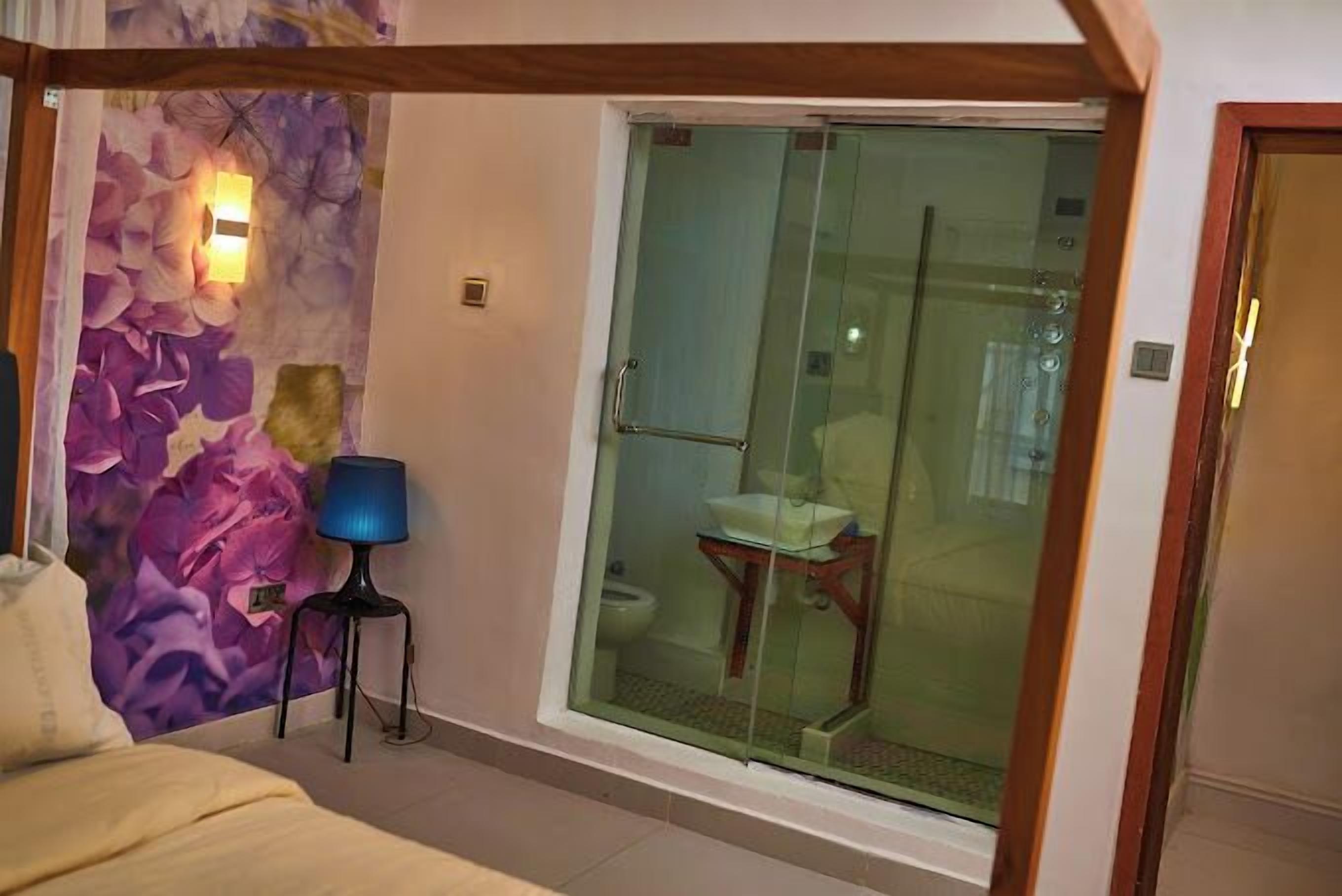 Een slaapkamer met een bed, nachtkastje, lamp, een glazen deur naar een badkamer en een wand met een bloemenmural.