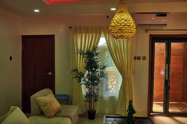 A living room with a sofa, a potted plant, a hanging lamp, and a sliding glass door leading to an outdoor area.