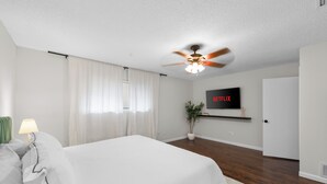A bedroom with a bed, a ceiling fan, a TV mounted on the wall, and a potted plant.
