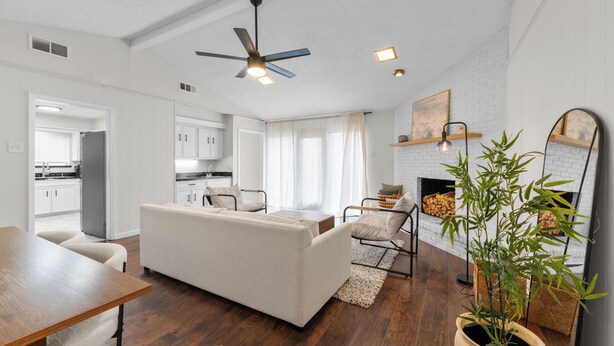 Un salon moderne avec un canapé, une table à manger et un ventilateur de plafond. On aperçoit au fond une cuisine avec des meubles blancs et une fenêtre.
