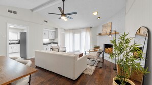 A modern living room with a sofa, a dining table, and a ceiling fan. There is a kitchen in the background with white cabinets and a window.